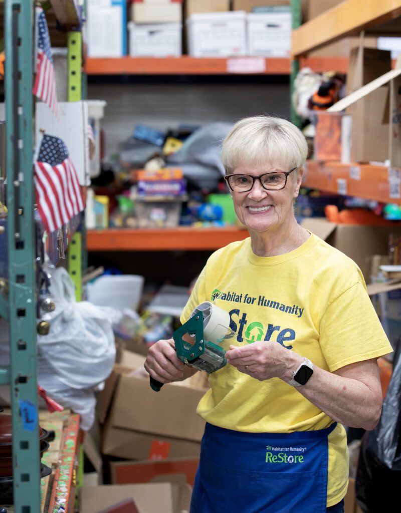 Debbie Hill, a senior volunteer at the Williamsburg Habitat ReStore, smiling while organizing items in the back warehouse.