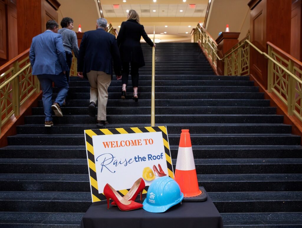 Raise the Roof welcome sign sitting at the bottom of the stairs with a pair of heels, a construction cone, and a hard hat. 