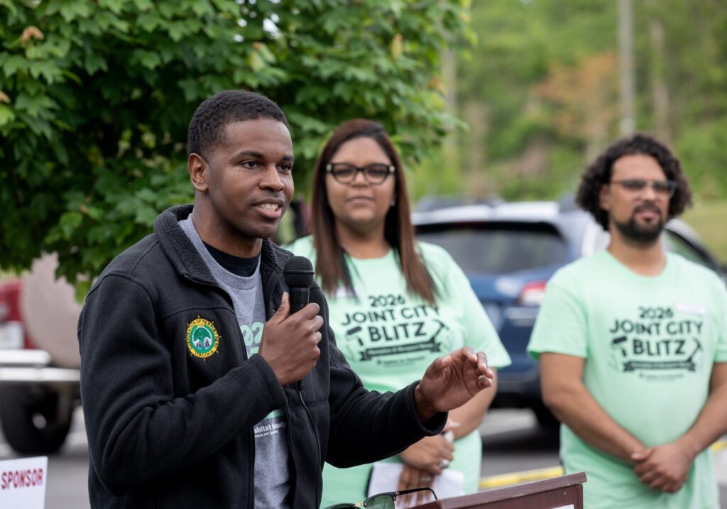 Man speaking into microphone and two people standing behind him watching