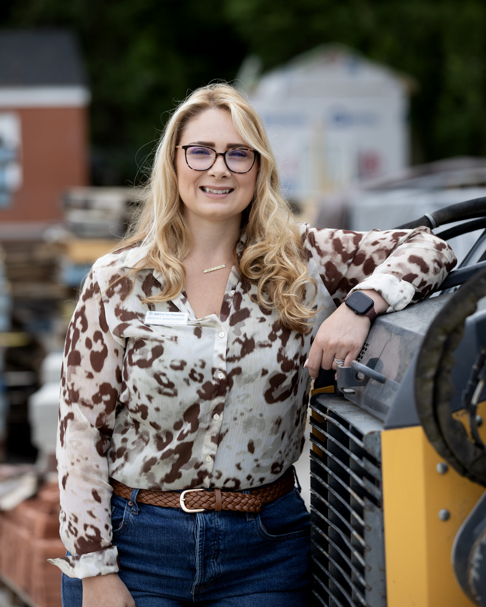 A woman with long blonde hair and glasses, wearing a patterned blouse and jeans, stands outdoors leaning on construction equipment.