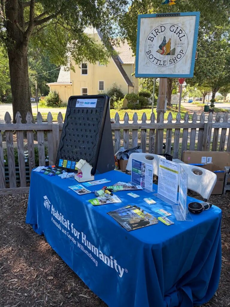 Habitat for Humanity info table at Bird Girl Bottle Shop, featuring brochures and a prize drop game under a sunny tree.

