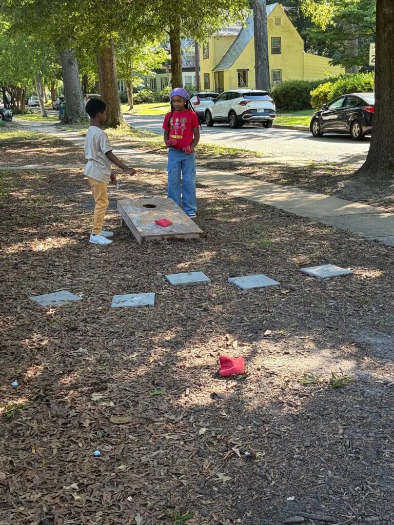 Two kids play cornhole on a leafy street, surrounded by trees and parked cars, with a yellow house in the background.