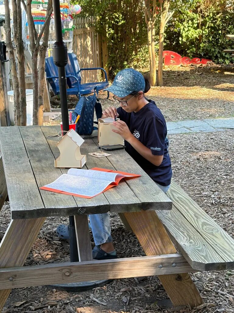 A person in a cap assembles a wooden model at a picnic table, with a coloring book and backpack nearby in a sunny garden.