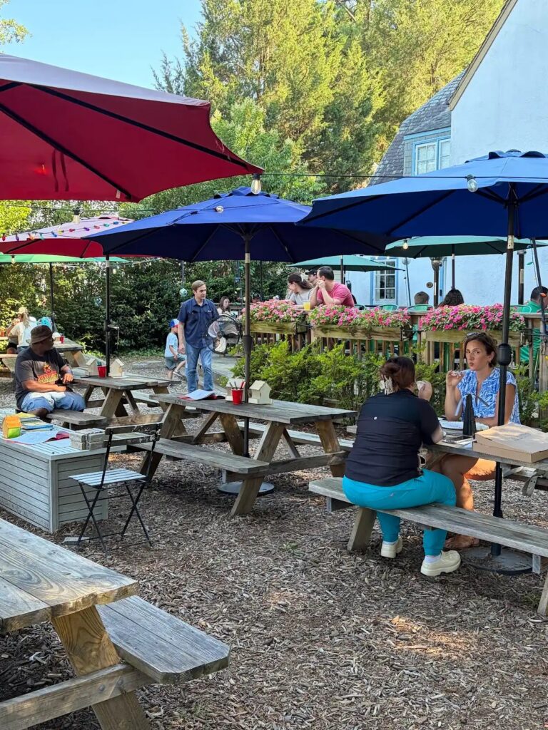People enjoy a sunny day at an outdoor cafe with picnic tables and colorful umbrellas, surrounded by greenery and flowers.