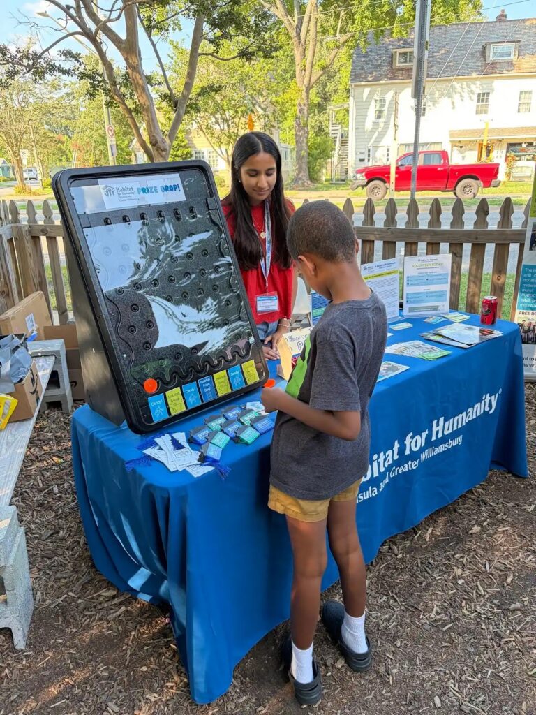 Child plays prize drop game at Habitat for Humanity booth outdoors, guided by a volunteer.