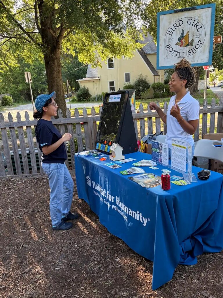 A Habitat for Humanity info table at Bird Girl Bottle Shop, with two people engaged in conversation under a tree.