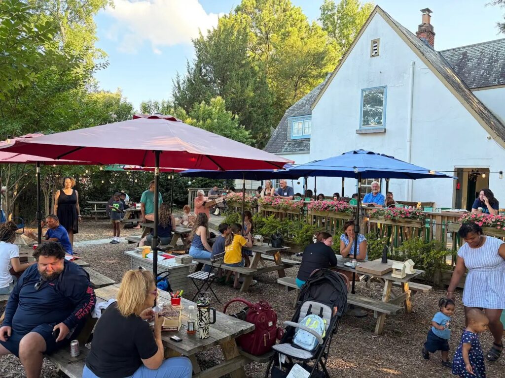 People enjoy a sunny day at a cafe garden with colorful umbrellas, surrounded by trees and a white house in the background.