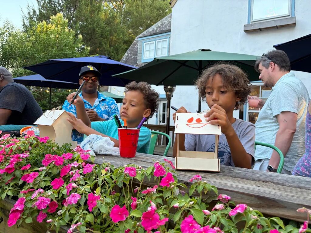 Children paint wooden boxes at an outdoor table, surrounded by pink flowers and adults, under large umbrellas.