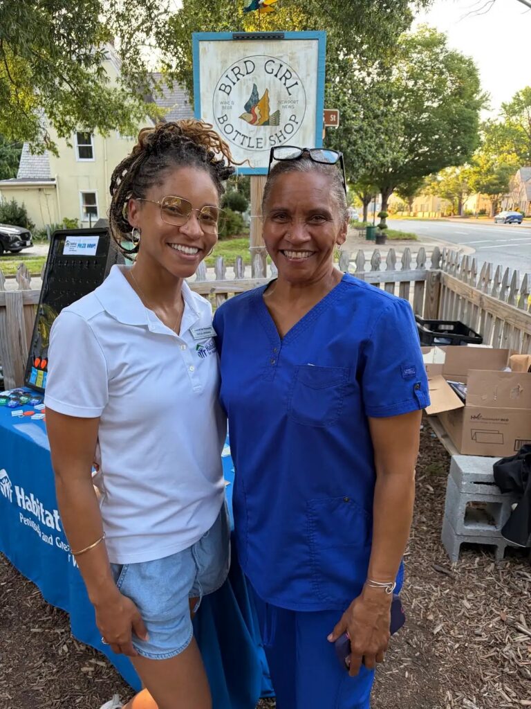 Two women smiling in front of Bird Girl Bottle Shop sign, one in a white Habitat shirt, the other in blue scrubs, outdoors.