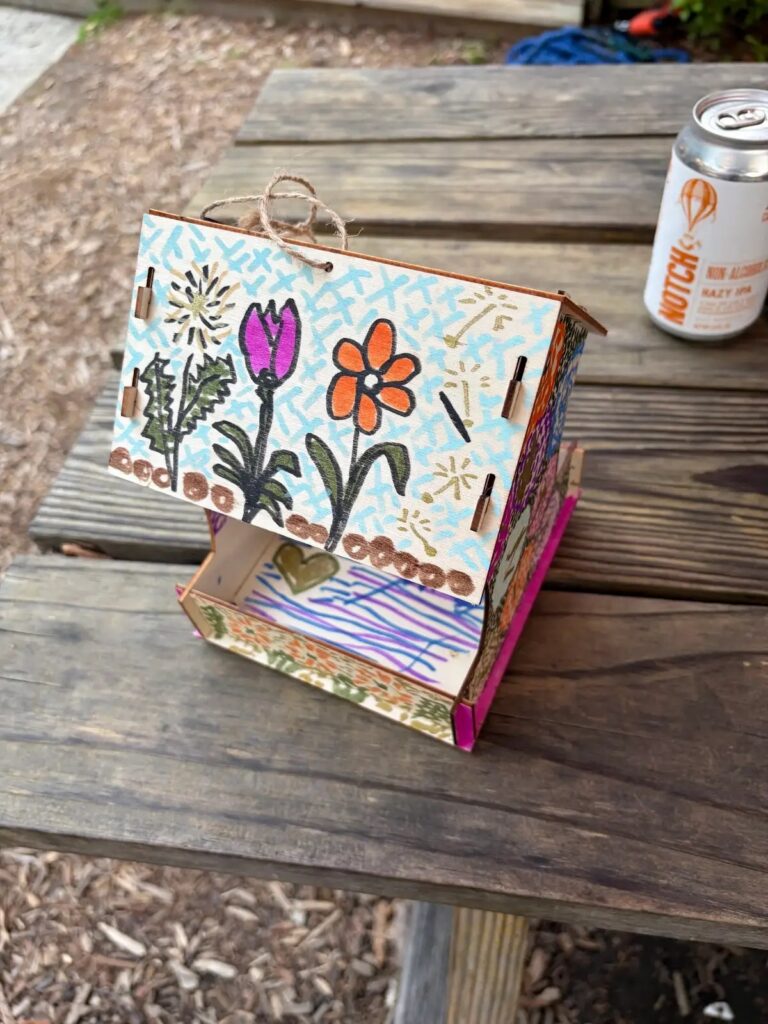 Colorful handmade birdhouse with floral designs on a wooden table, next to a can.