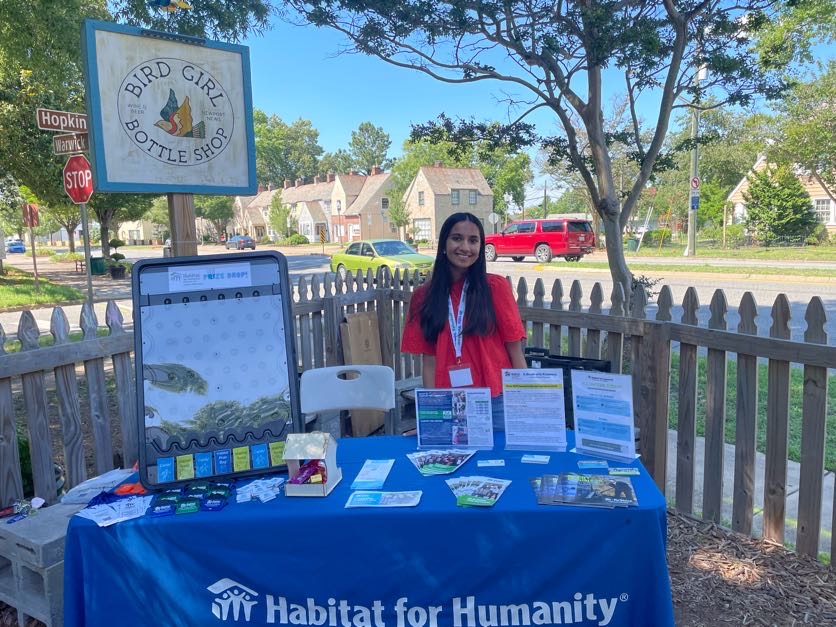 A woman stands by a Habitat for Humanity table with brochures, under a "Bird Girl Bottle Shop" sign, on a sunny street corner.