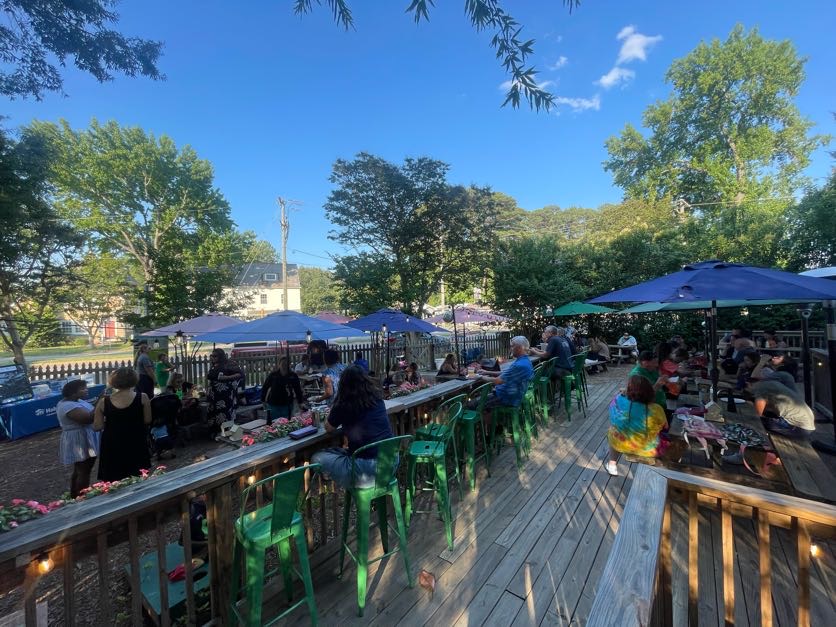 People enjoy a sunny day at an outdoor cafe with green chairs and blue umbrellas, surrounded by trees and a picket fence.
