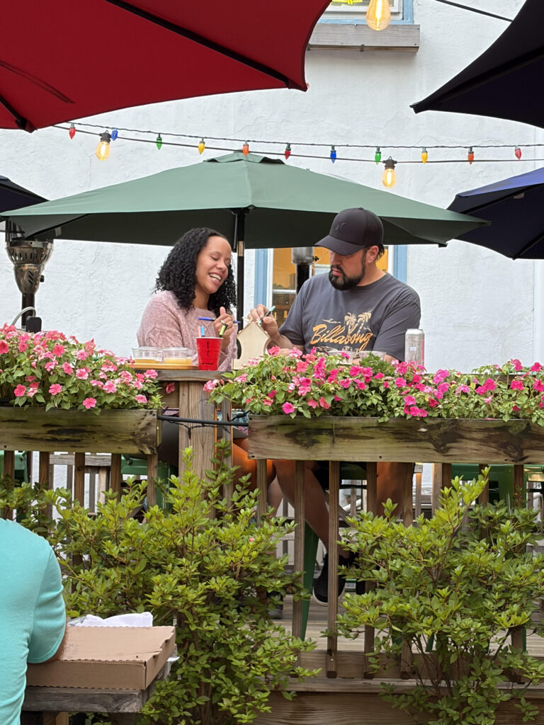A couple enjoys a meal at an outdoor cafe, surrounded by pink flowers and colorful lights under umbrellas.