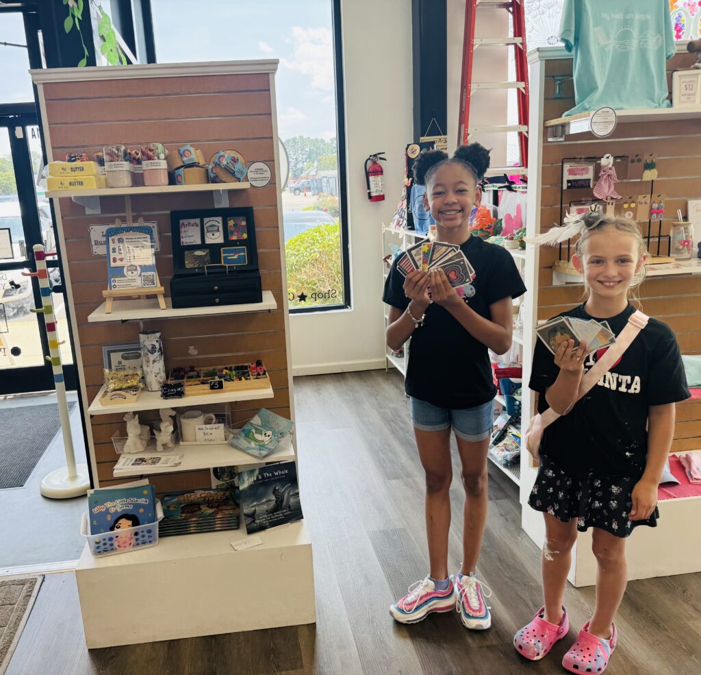 Two smiling girls hold cards in a colorful shop, standing beside shelves filled with toys and crafts, sunlight streaming in.