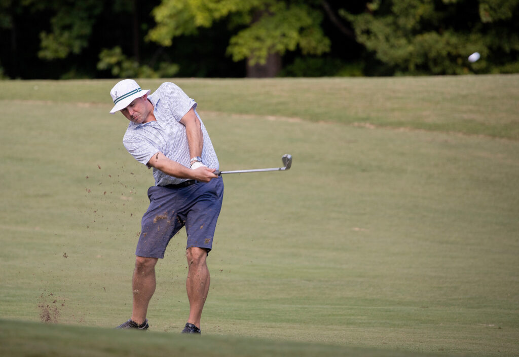A man in a hat swings a golf club on a grassy course, hitting a ball into the air.