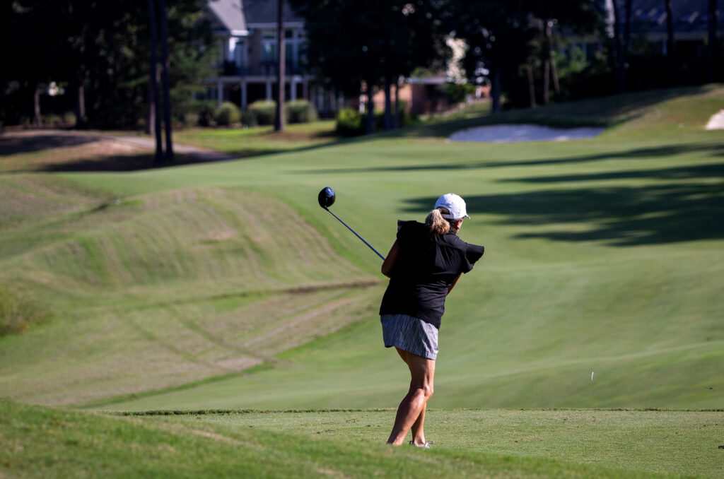 A woman swings a golf club on a sunny golf course with houses in the background.