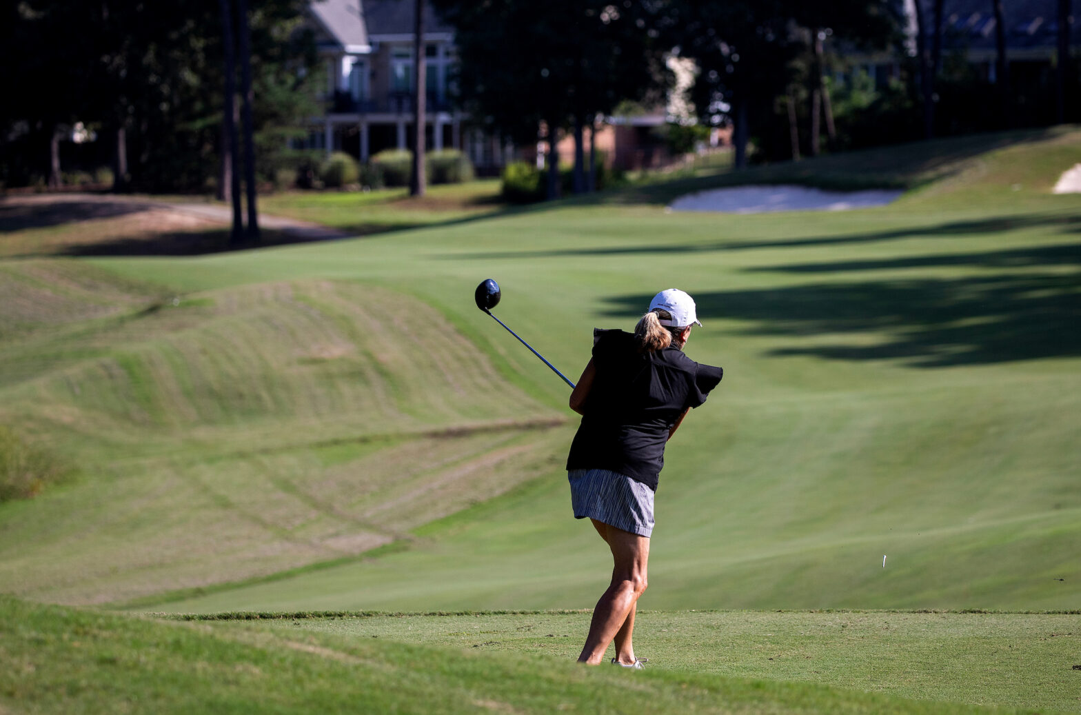 A woman swings a golf club on a sunny golf course with houses in the background.