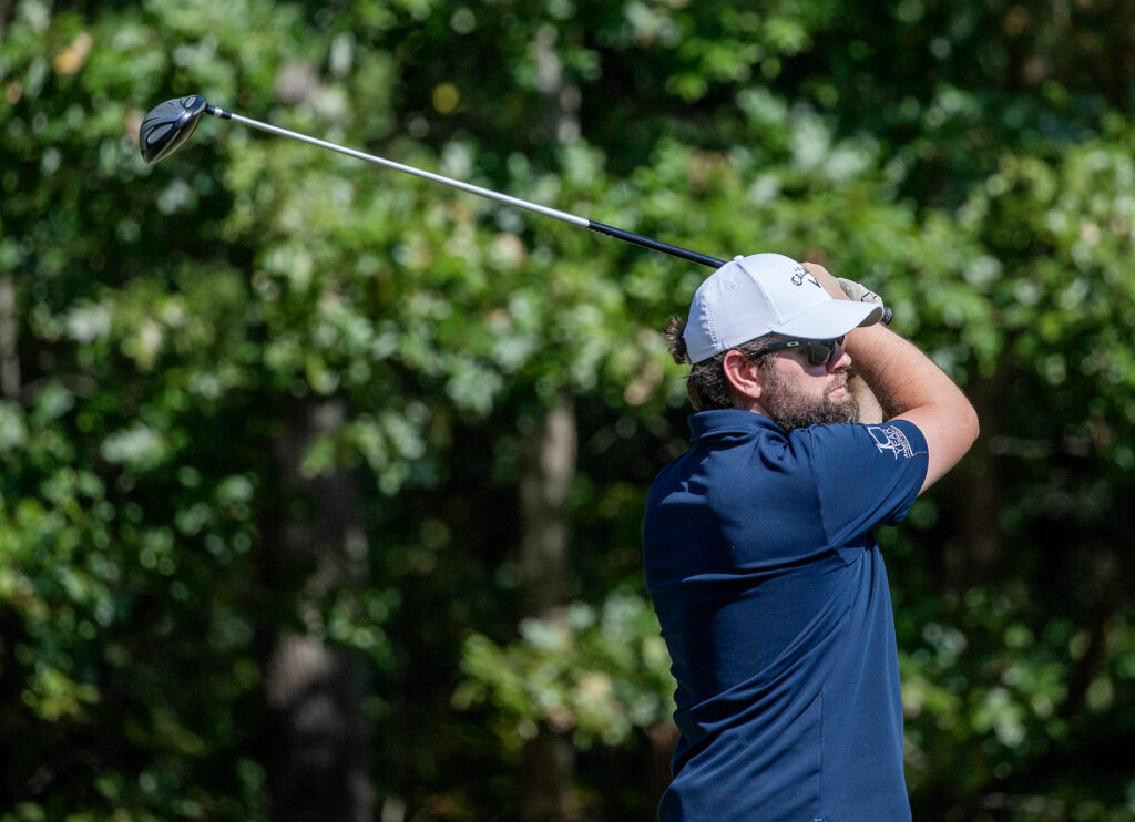 Golfer in navy shirt and white hat follows through on a swing, green trees in the background.