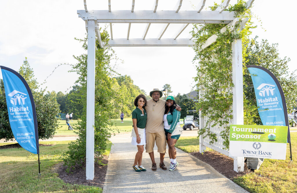 Three people pose under a white pergola at a golf event with Habitat for Humanity banners on each side.