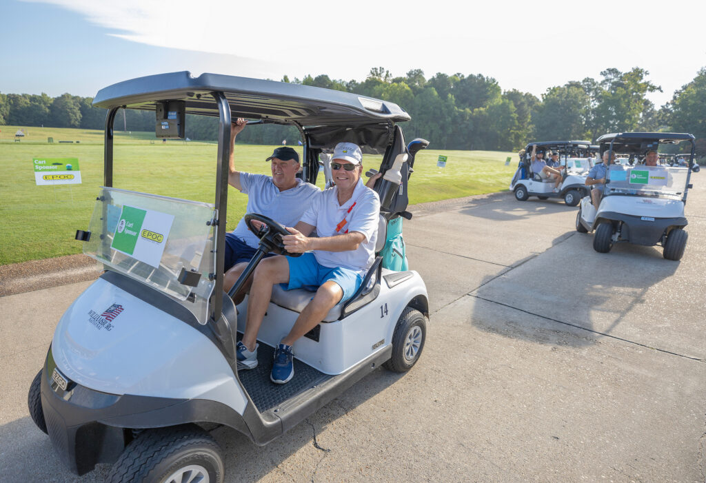 Two men in a golf cart smile at the camera, with other carts and people visible in the background.