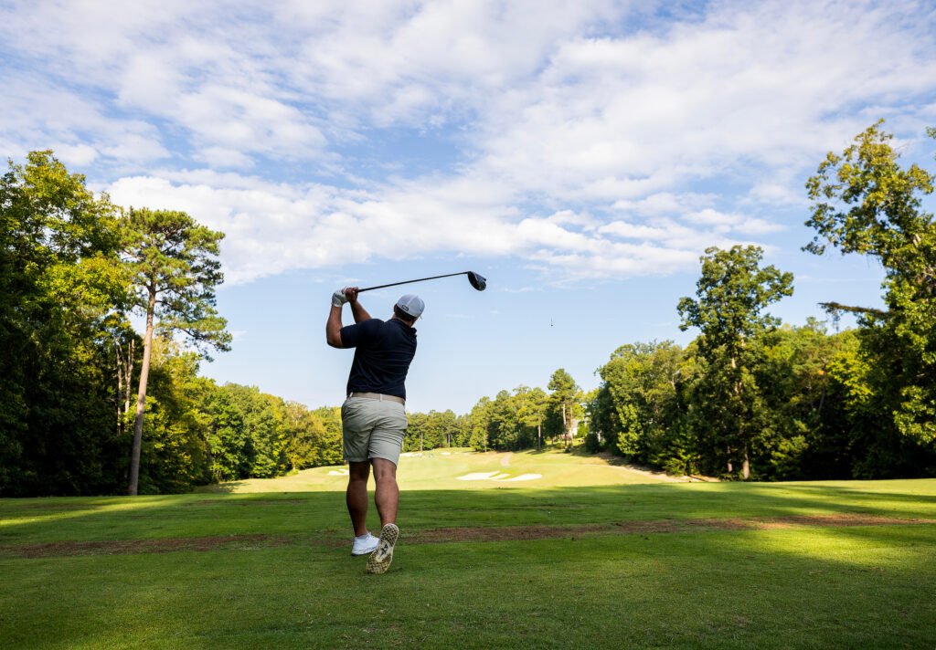 A person in shorts and a cap swings a golf club on a sunny course lined with trees.