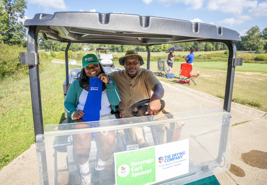 Two people sit in a golf cart on a sunny course, with sponsor signs on the windshield and others behind them.