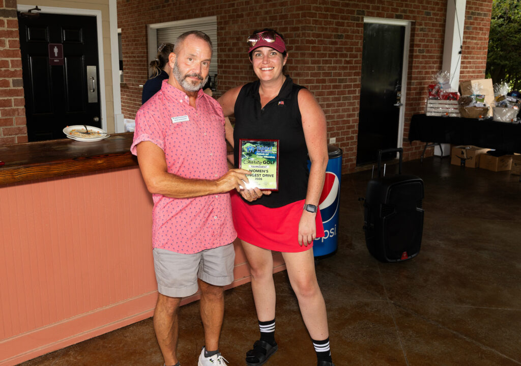 Two people pose indoors, one holding a plaque that reads “2023 Golf Champion’s Closet Drive.”.