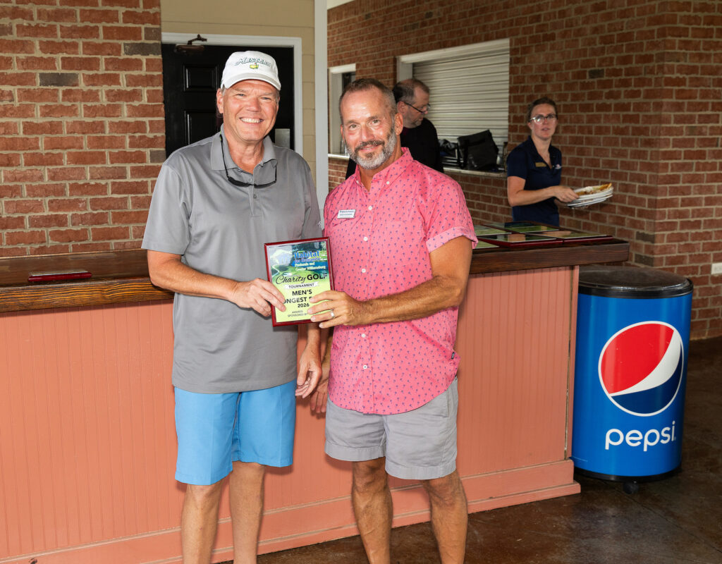 Two men smiling and holding a “Men’s Flight” plaque, standing indoors by a counter with a Pepsi cooler.