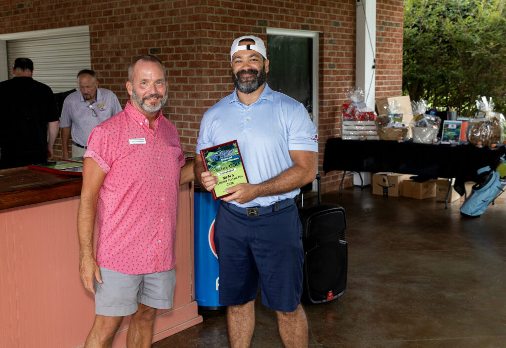 Two men pose indoors; one holds a plaque. Gift baskets and boxes are on a table in the background.